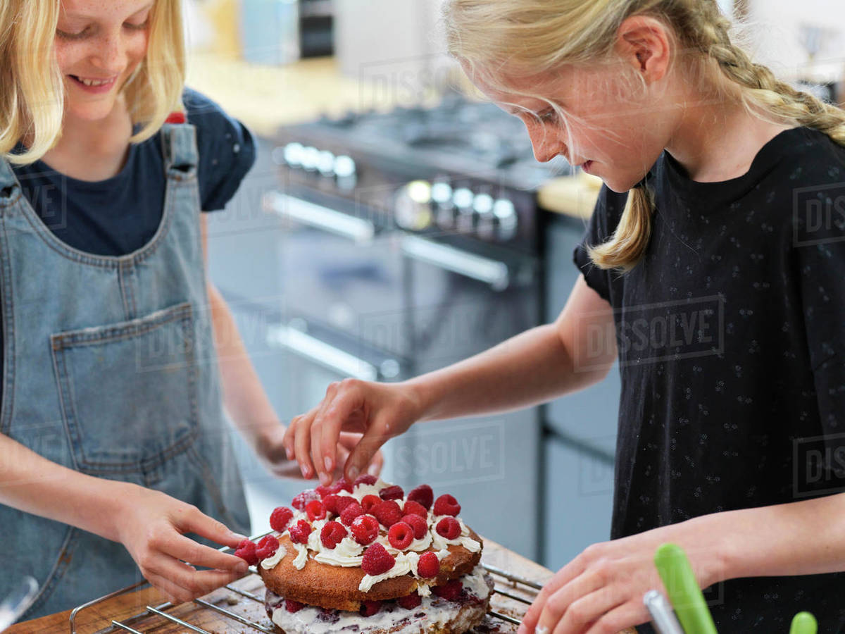 Girl and her sister baking a cake, decorating cake with fresh cream and raspberries at kitchen table Royalty-free stock photo