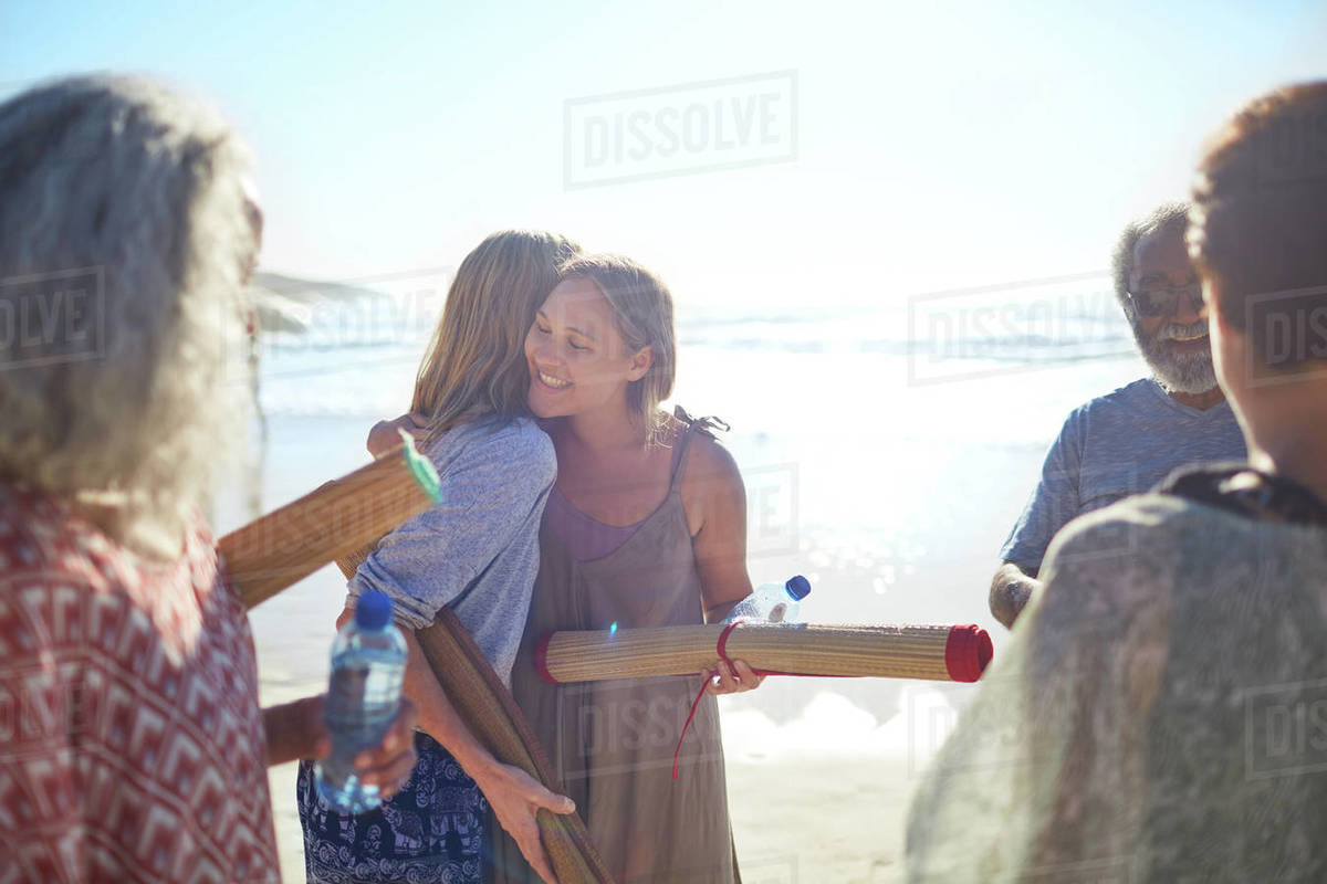 Women friends with yoga mats hugging on sunny beach during yoga retreat