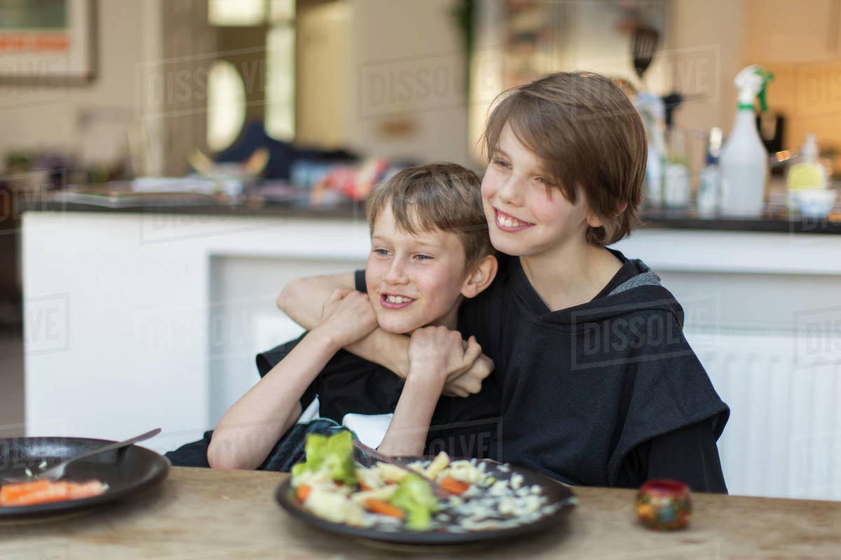 Happy brothers hugging and eating dinner at dining table Stock Photo