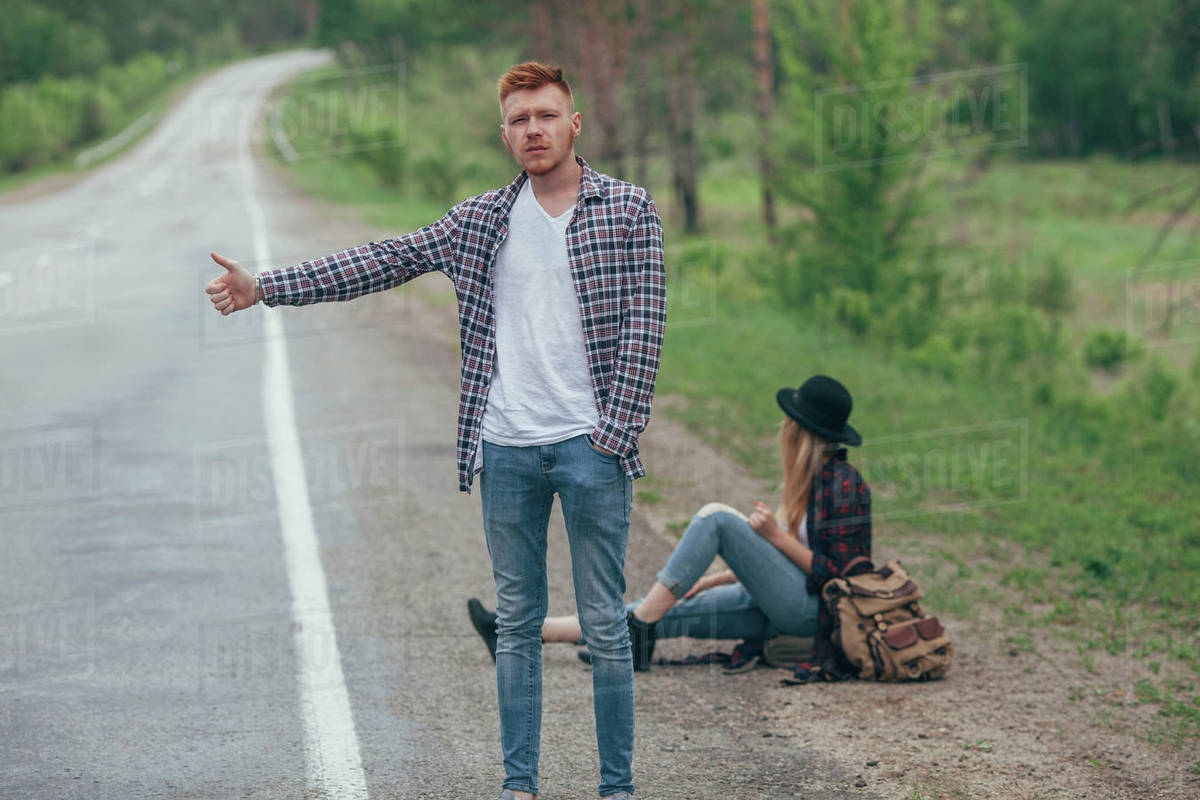 Man hitchhiking while woman sitting on roadside Stock Photo Dissolve